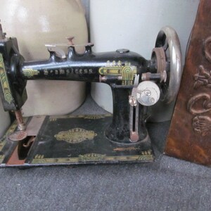 Op de afbeelding: Een zwarte vintage naaimachine met gouden accenten en de woorden "The " en "Sew-Princess" aan de zijkant. De machine heeft een voetpedaal en een handwiel.