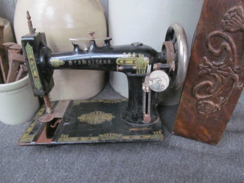 Op de afbeelding: Een zwarte vintage naaimachine met gouden accenten en de woorden "The " en "Sew-Princess" aan de zijkant. De machine heeft een voetpedaal en een handwiel.