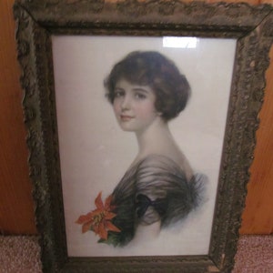May include: A framed vintage print of a woman with dark hair wearing a black dress and holding a red poinsettia flower. The frame is ornate with a gold finish.
