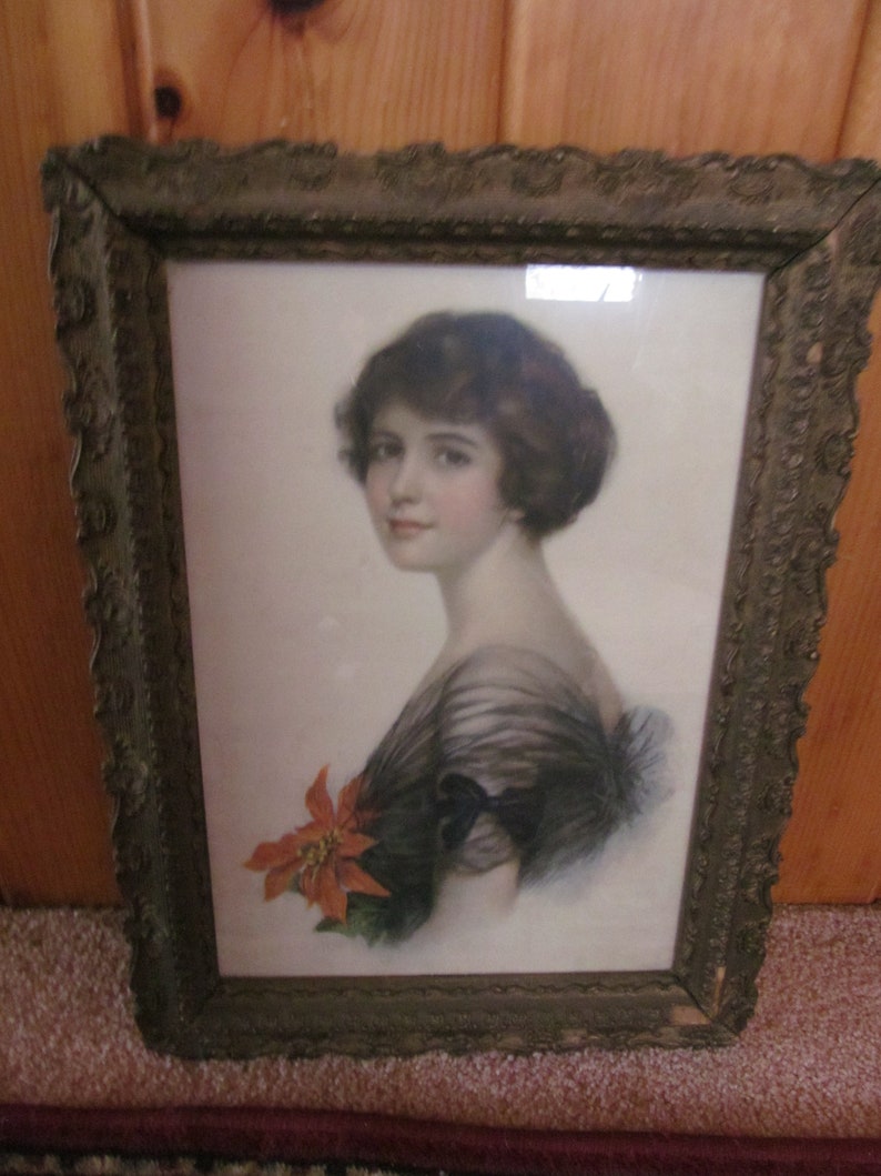 May include: A framed vintage print of a woman with dark hair wearing a black dress and holding a red poinsettia flower. The frame is ornate with a gold finish.