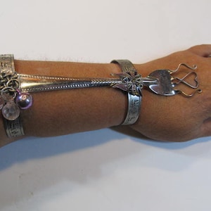 Puede incluir: Un brazalete de plata con un tenedor unido a él. El tenedor se ha doblado en forma de corazón. El brazalete tiene cuentas y abalorios rosados.