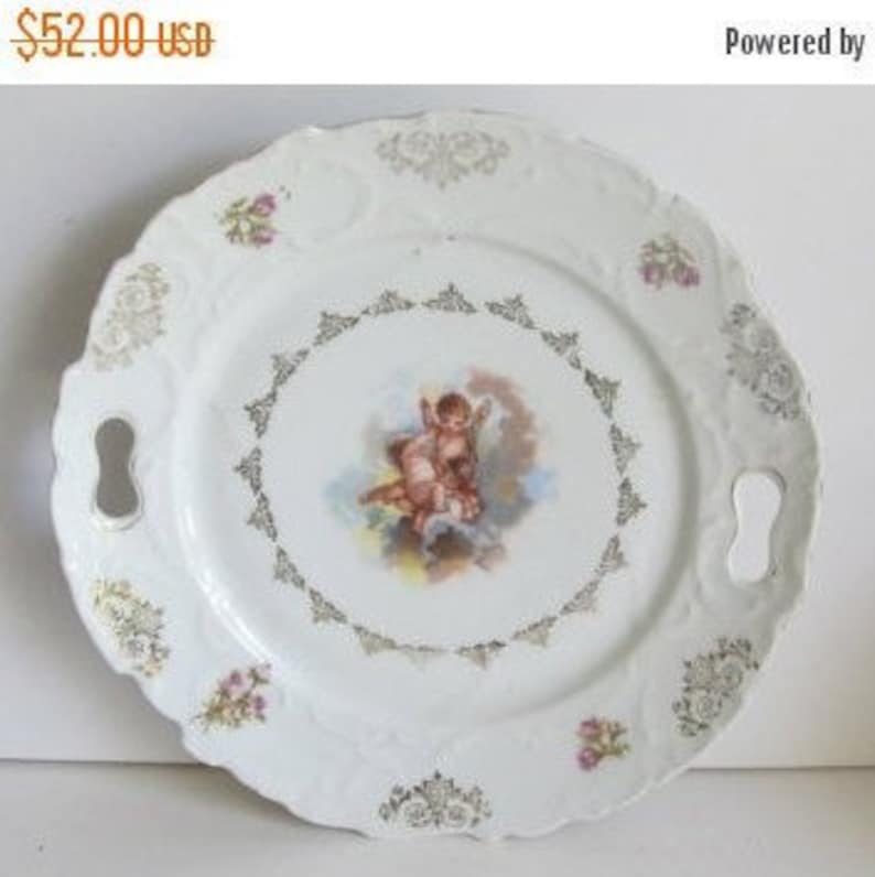 May include: White porcelain serving plate with gold trim and a cherub design. The plate has two handles.