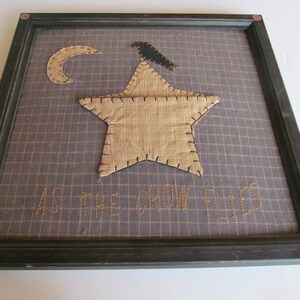 May include: Framed artwork featuring a tan star and crescent moon applique on a grid background. A black crow sits atop the star. The phrase "AS THE CROW FLIES" is stitched below the star in gold thread. The frame is dark green.