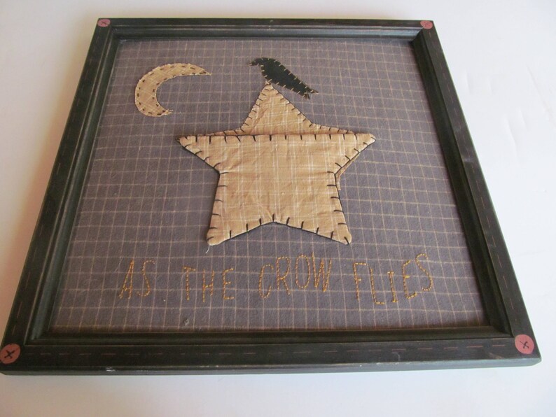 May include: Framed artwork featuring a tan star and crescent moon applique on a grid background. A black crow sits atop the star. The phrase "AS THE CROW FLIES" is stitched below the star in gold thread. The frame is dark green.