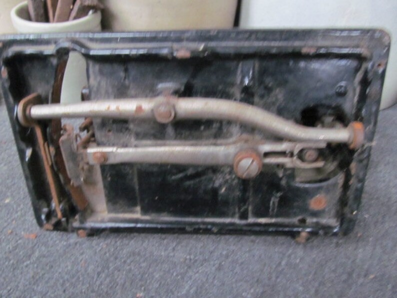 Op de afbeelding: Close-up van de onderkant van een vintage zwarte naaimachine met een zilveren metalen hendel en een zilveren metalen arm.