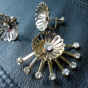 Pode incluir: Um par de brincos de press&atilde;o dourados com strass transparentes. Os brincos t&ecirc;m a forma de uma flor com uma pedra central e pedras menores ao redor das bordas.