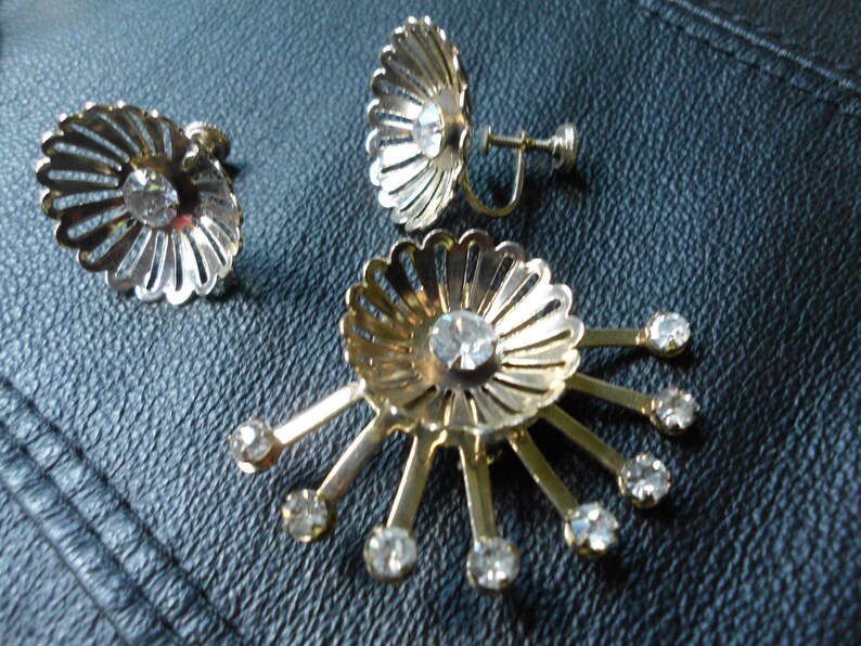 Pode incluir: Um par de brincos de press&atilde;o dourados com strass transparentes. Os brincos t&ecirc;m a forma de uma flor com uma pedra central e pedras menores ao redor das bordas.