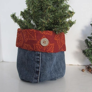 Puede incluir: Una maceta de mezclilla con un forro de tela rojo y naranja y un botón. La maceta está llena de un pequeño árbol de hoja perenne artificial.