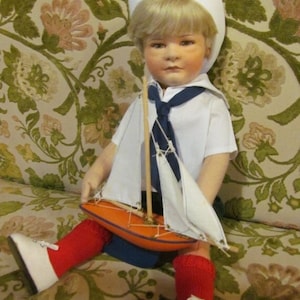 Op de afbeelding: Een vintage pop gekleed in een matrozenoutfit, compleet met een witte hoed, overhemd en marineblauwe stropdas. De pop houdt een kleine oranje zeilboot met witte zeilen vast. De pop draagt rode sokken en witte schoenen.