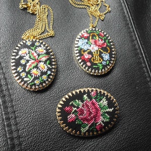 May include: Three oval-shaped embroidered pendants with floral designs. The pendants are on gold chains and have black fabric with colorful embroidery. One pendant features a red flower with green leaves, another has a pink rose with green leaves, and the third has a white flower with red and green leaves.