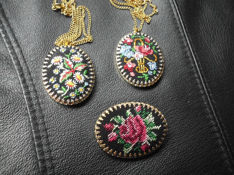 May include: Three oval-shaped embroidered pendants with floral designs. The pendants are on gold chains and have black fabric with colorful embroidery. One pendant features a red flower with green leaves, another has a pink rose with green leaves, and the third has a white flower with red and green leaves.