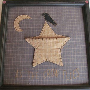 May include: Framed textile art featuring a tan star with a black crow perched on top, a crescent moon, and the embroidered phrase "AS THE CROW FLIES." The background is a grid pattern in shades of gray and tan.
