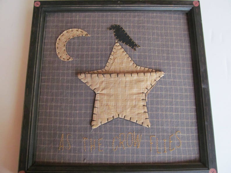 May include: Framed textile art featuring a tan star with a black crow perched on top, a crescent moon, and the embroidered phrase "AS THE CROW FLIES." The background is a grid pattern in shades of gray and tan.
