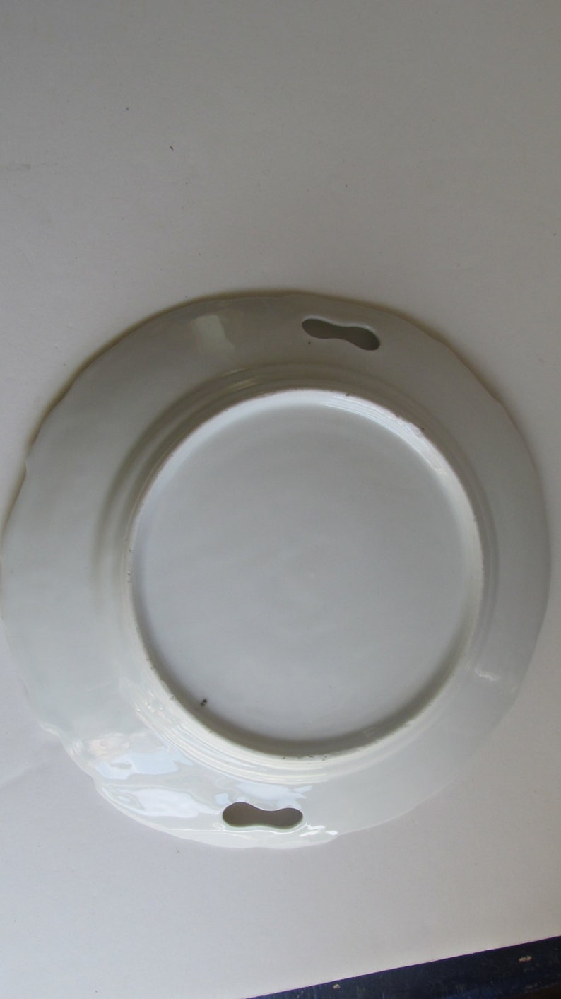 May include: White ceramic plate with a scalloped edge and two holes on either side. The plate is designed for serving food.