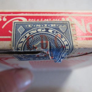 May include: A vintage playing card deck box with a red and white label. The label has the text "REG. U.S. PAT. OFF." and "No. 9". The box has a blue and white seal with the text "Playing Cards" and a diamond and heart symbol.