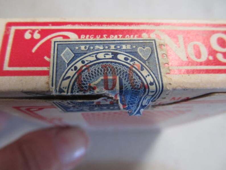 May include: A vintage playing card deck box with a red and white label. The label has the text "REG. U.S. PAT. OFF." and "No. 9". The box has a blue and white seal with the text "Playing Cards" and a diamond and heart symbol.