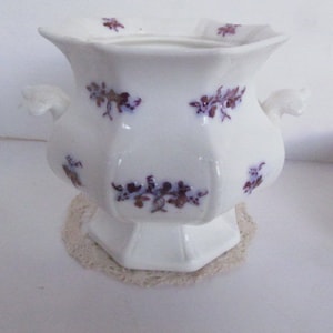 Può includere: Una zuccheriera in porcellana bianca con un bordo ondulato e due manici. La ciotola è decorata con un motivo floreale viola.