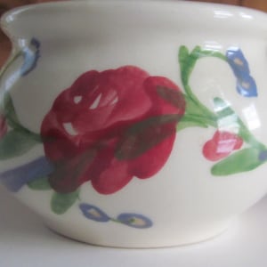 Può includere: Una ciotola in ceramica bianca con un motivo floreale. La ciotola presenta una grande rosa rossa con foglie verdi e fiori blu.