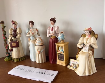 May include: Five porcelain figurines of women dressed in vintage clothing. Each figurine is holding a different object, including a suitcase, a cake, a globe, a book, and a sewing kit. The figurines are all standing on a wooden surface.