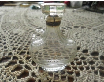 May include: A clear glass bottle with a gold stopper and the words "L'Air du Temps" written on the front. The bottle is sitting on a white crocheted surface.