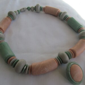 May include: A necklace made of pastel pink and green ceramic beads. The necklace is made of a variety of shapes and sizes of beads. There is a matching pendant with a pink ceramic bead.