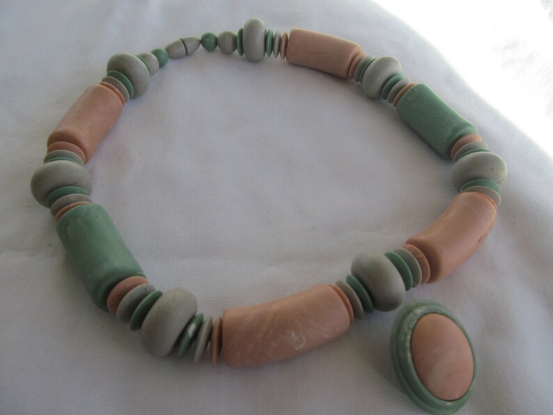 May include: A necklace made of pastel pink and green ceramic beads. The necklace is made of a variety of shapes and sizes of beads. There is a matching pendant with a pink ceramic bead.