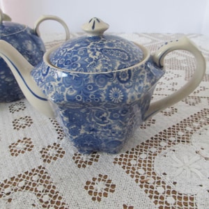 May include: A blue and white teapot with a floral pattern. The teapot has a lid and a handle. The teapot is sitting on a white lace tablecloth.