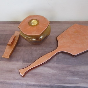 May include: A vintage vanity set with a peach-colored glass jar with a gold lid, a matching peach-colored hand mirror, and a small peach-colored tray with a handle.