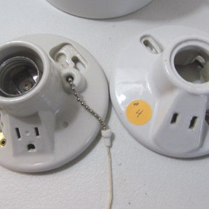 May include: Two white ceramic light fixture bases with a light bulb socket and a built-in electrical outlet. The base on the left has a pull chain. The base on the right has the number "4" written on it.