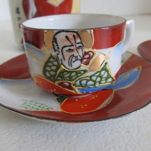 May include: A red and white teacup and saucer set with a hand-painted design of a man's face with a gold trim. The design includes green, blue, and gold accents.