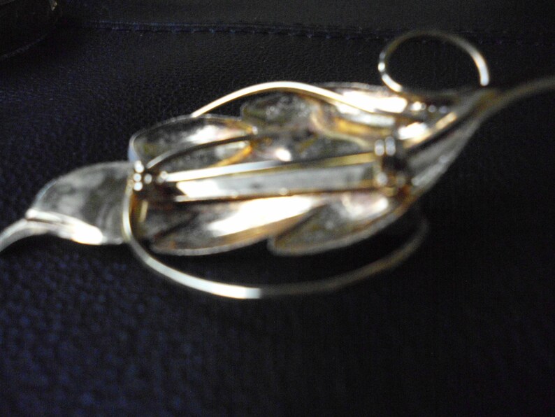 May include: A gold-toned leaf-shaped brooch with a pin closure on the back. The brooch has a textured surface and a delicate design.