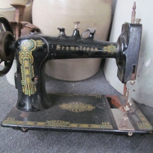 Op de afbeelding: Een zwarte vintage naaimachine met een gouden en groen decoratief ontwerp. De machine heeft een handwiel en een naaldplaat. De merknaam "Seamsstress" is zichtbaar op de machine.