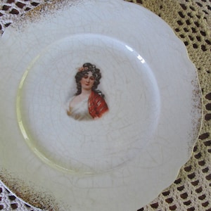 May include: A white porcelain plate with a gold rim and a painted image of a woman in a white dress and red shawl.