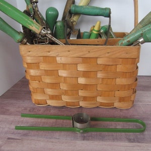 Puede incluir: Una cesta de mimbre llena de herramientas de jardiner&iacute;a, incluyendo una azada verde, un rastrillo de mano verde y un cultivador de mano verde. Las herramientas est&aacute;n dispuestas en la cesta con sus mangos sobresaliendo. La cesta est&aacute; sentada sobre una superficie de madera.