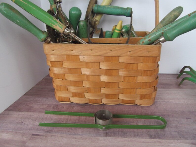 Puede incluir: Una cesta de mimbre llena de herramientas de jardiner&iacute;a, incluyendo una azada verde, un rastrillo de mano verde y un cultivador de mano verde. Las herramientas est&aacute;n dispuestas en la cesta con sus mangos sobresaliendo. La cesta est&aacute; sentada sobre una superficie de madera.