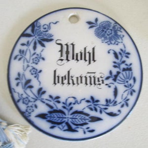 May include: A white oval ceramic ornament with blue floral designs and the text "Willkommen" in black script.