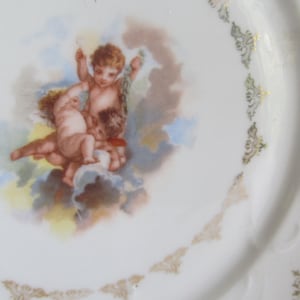 May include: A white porcelain plate with a gold trim and a central image of three cherubs in a cloud-like setting. The plate has a scalloped edge and a handle on the side.