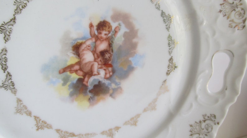 May include: A white porcelain plate with a gold trim and a central image of three cherubs in a cloud-like setting. The plate has a scalloped edge and a handle on the side.
