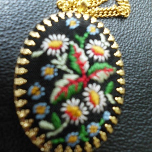 May include: A gold chain necklace with a black oval pendant featuring embroidered flowers in shades of red, white, blue, and green.