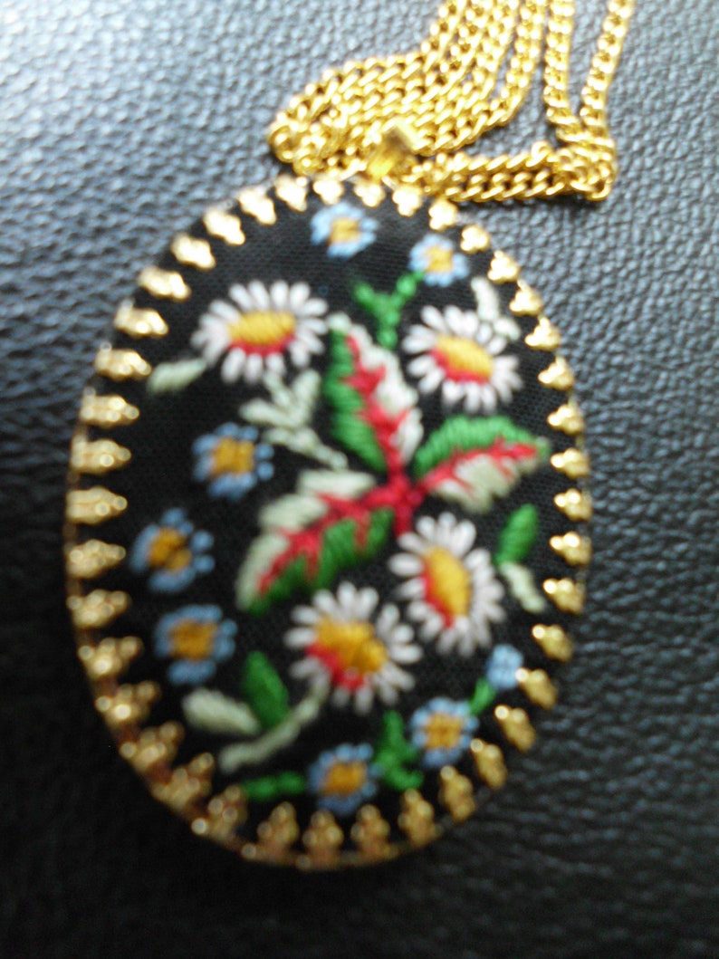 May include: A gold chain necklace with a black oval pendant featuring embroidered flowers in shades of red, white, blue, and green.