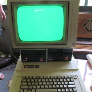 以下が含まれることがあります： 緑色の画面に「Apple IIe」というテキストが表示された、ビンテージのApple IIeコンピューター。コンピューターはベージュのケースで、黒いキーボードと「disk II」というラベルの付いたディスクドライブが2つあります。