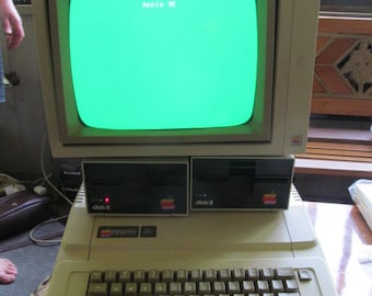 May include: A vintage Apple IIe computer with a green screen displaying the text "Apple IIe". The computer has a beige case with a black keyboard and two disk drives labelled "disk II".