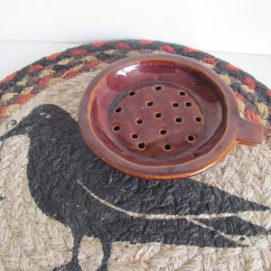 May include: A brown ceramic dish with a handle and holes in the bottom. The dish is sitting on a woven rug with a black crow design.
