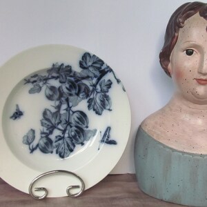 Può includere: Un piatto in ceramica bianca con un design floreale blu e bianco che presenta un ramo di ribes e un'ape. Il piatto è su una superficie di legno accanto a un busto in ceramica di una donna con un vestito blu.