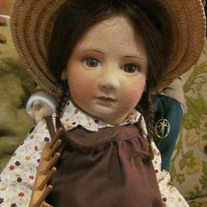 May include: A brown-haired doll wearing a brown dress with a white floral pattern, a straw hat, and a wooden rake. The doll has brown eyes and a light complexion.