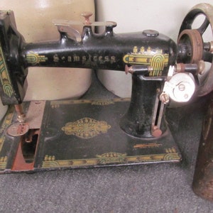 Op de afbeelding: Antiek zwarte naaimachine met gouden en groene decoratieve accenten. De machine heeft een handwiel en een voetpedaal. De merknaam "Scramstress" is op de machine gedrukt.