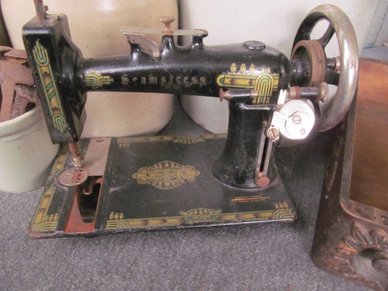 Op de afbeelding: Antiek zwarte naaimachine met gouden en groene decoratieve accenten. De machine heeft een handwiel en een voetpedaal. De merknaam "Scramstress" is op de machine gedrukt.