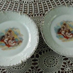 May include: Two white porcelain plates with gold trim and a central image of cherubs in a cloud-like setting. The plates have a scalloped edge and are perfect for displaying or serving.