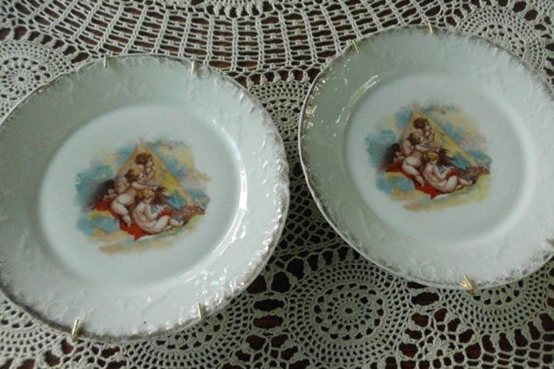 May include: Two white porcelain plates with gold trim and a central image of cherubs in a cloud-like setting. The plates have a scalloped edge and are perfect for displaying or serving.