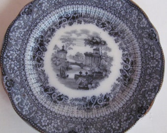 Puede incluir: Un plato de transferencia azul y blanco con una escena en blanco y negro de un puente y edificios. El plato tiene un borde festoneado y un diseño floral.